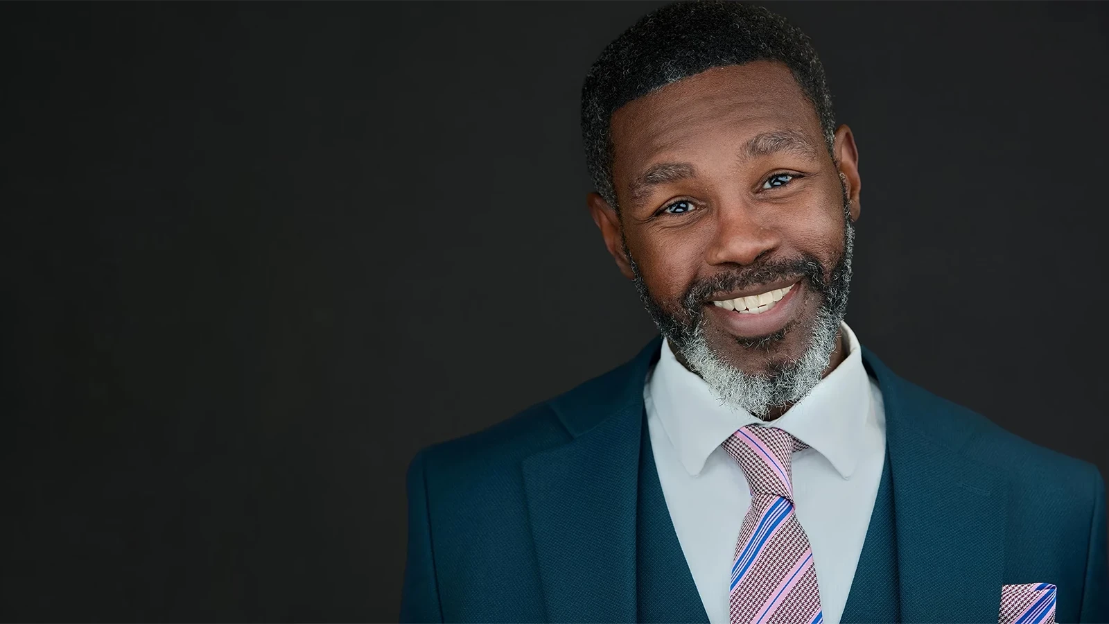 Professional headshot of a man in a teal suit and striped tie, broad natural smile