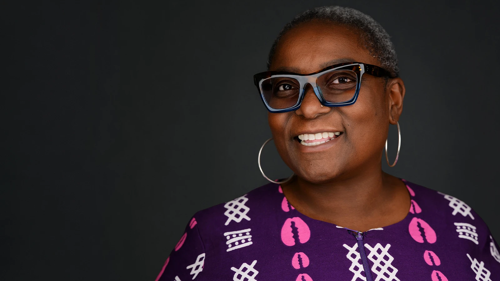 Professional headshot of a woman with short grey hair and blue-rimmed glasses in a purple patterned top, bright natural smile