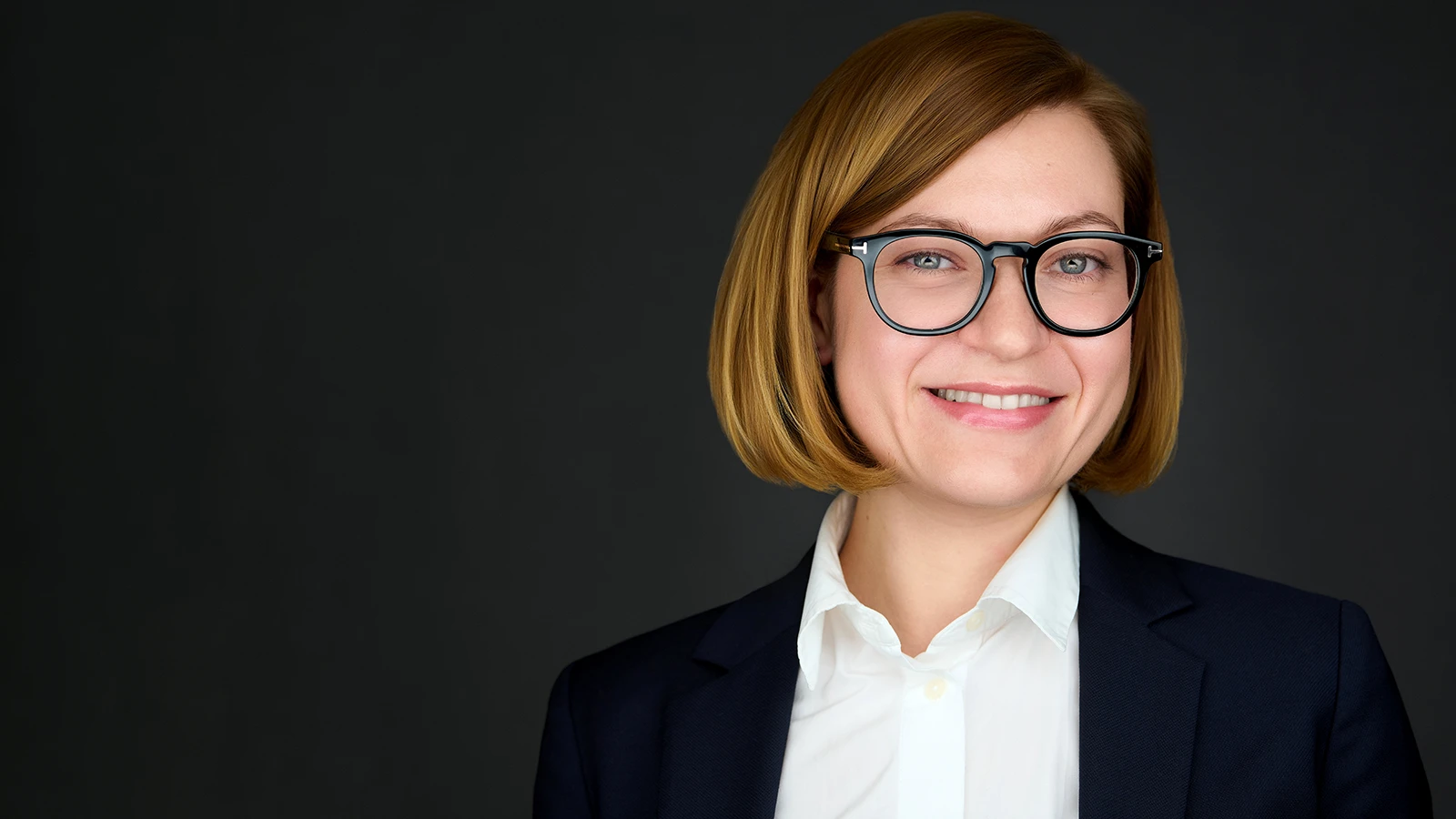 Corporate headshot of a woman in a navy blazer and glasses, photographed against a dark studio background