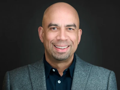Man in blazer and dress shirt, calm professional expression against a dark studio background