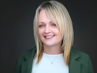 Woman in a dark blazer and blouse, confident and natural expression against a dark background