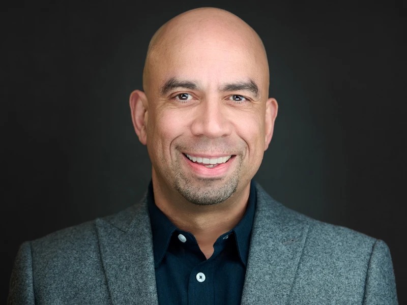 Professional headshot of a bald man with a goatee in a grey textured blazer, broad warm smile