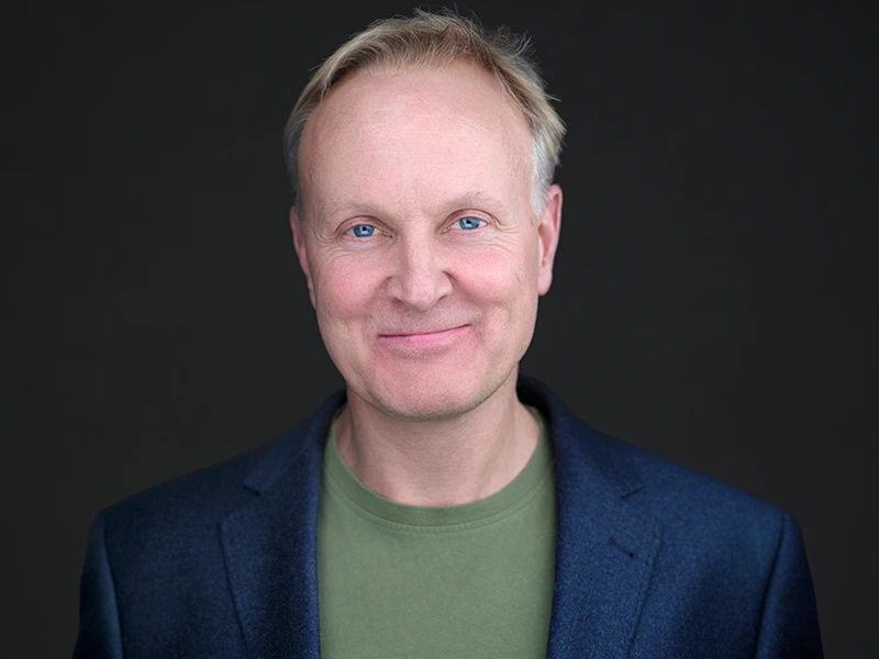 Professional headshot of a man in a navy blazer over a green jumper, relaxed approachable expression