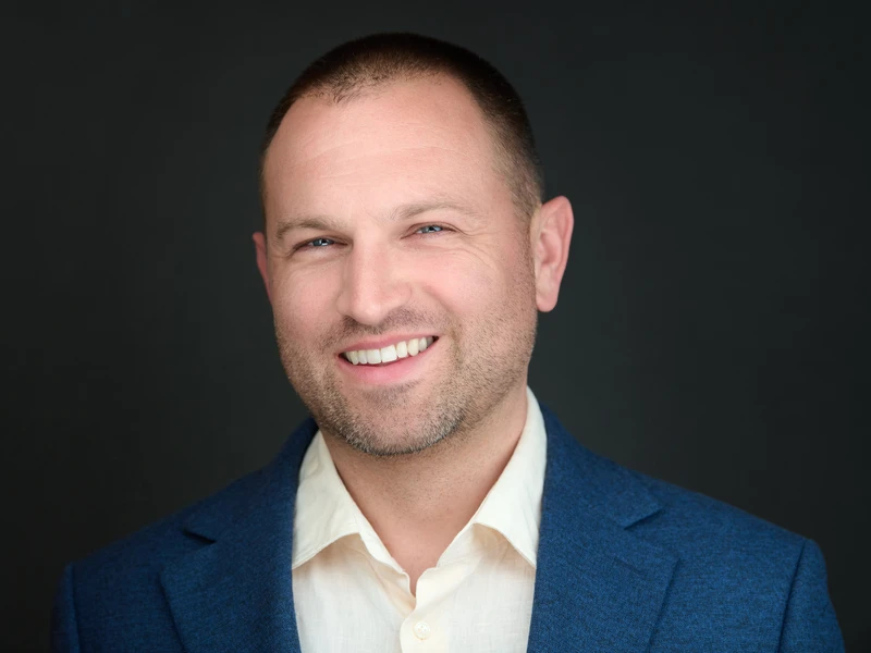 Professional headshot of a man in a navy blazer and open-collar shirt, warm confident smile