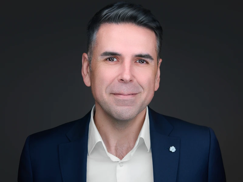 Professional headshot of a man with dark hair in a navy blazer and open-collar shirt, composed expression