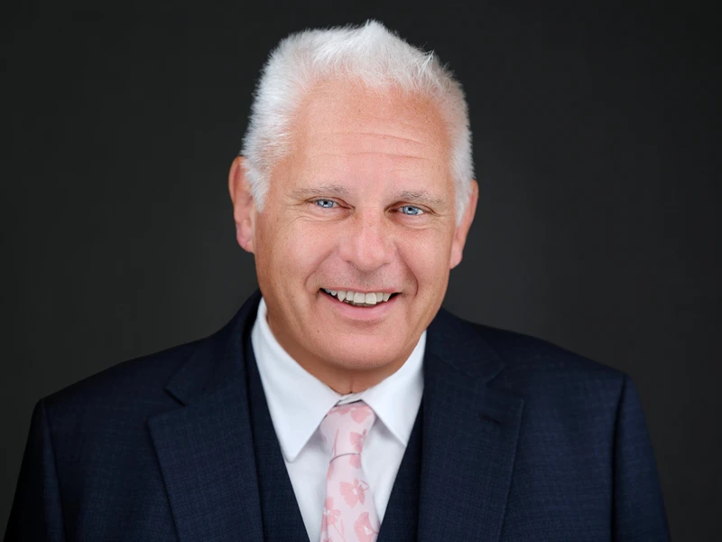 Professional business headshot of a smiling man with silver hair in a navy three-piece suit and pink tie on a dark grey background