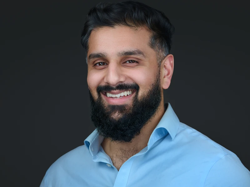 Professional headshot of a bearded man in a light blue open-collar shirt, broad natural smile