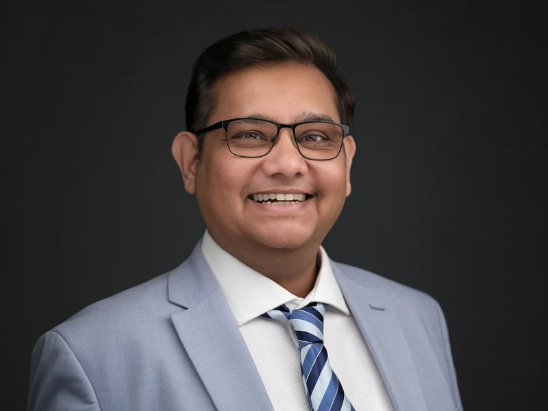 Corporate headshot of a man in glasses wearing a dark suit and shirt, calm professional expression on a dark studio background