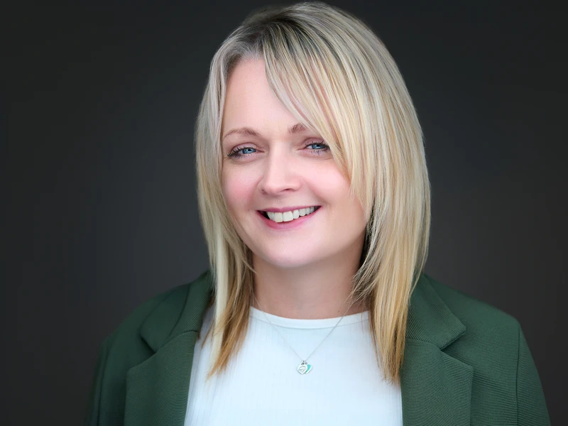 Professional headshot of a blonde woman in a dark green blazer, warm natural smile