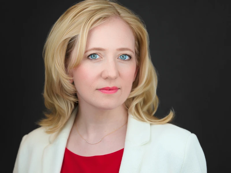 Corporate headshot of a woman in a white blazer over a red top, composed natural expression, dark studio background