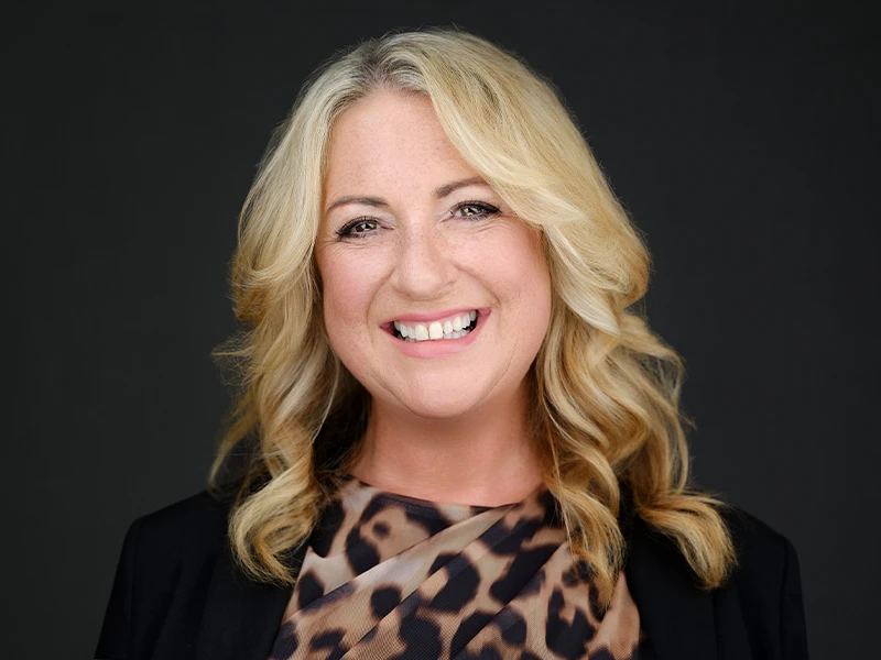 Business headshot of a smiling blonde woman in a black blazer over an animal-print top on a dark grey background