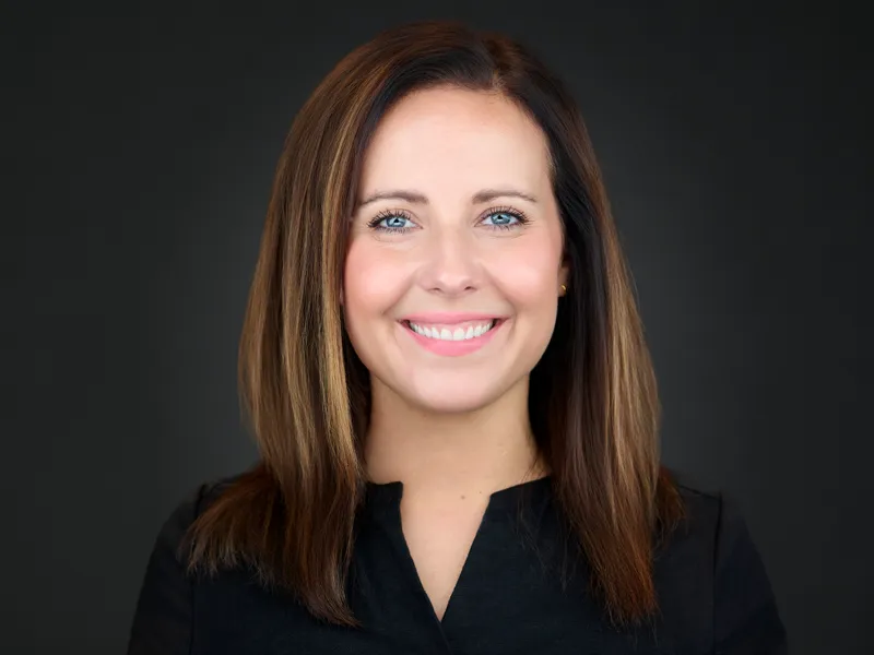 Professional headshot of a woman with long brown hair in a black blouse, bright natural smile