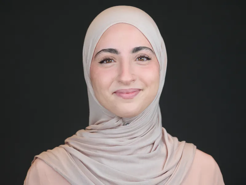 Professional headshot of a woman wearing a hijab, warm approachable smile, dark studio background
