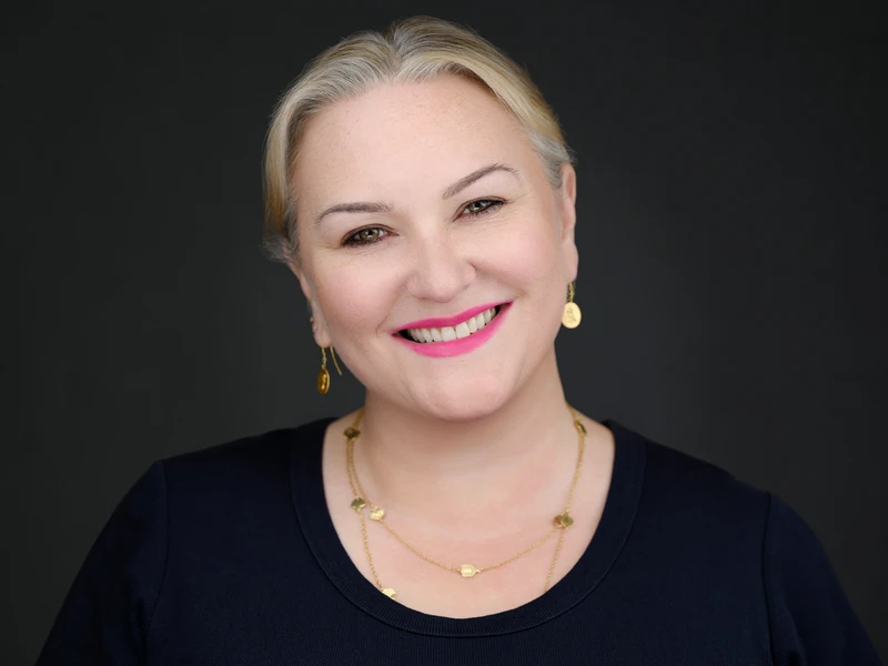 Business headshot of a smiling blonde woman in a navy top with a gold necklace on a dark grey background