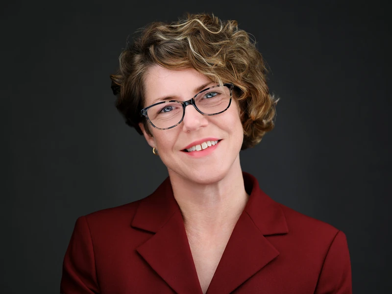 Business headshot of a smiling woman with short curly hair and glasses in a red blazer on a dark grey background