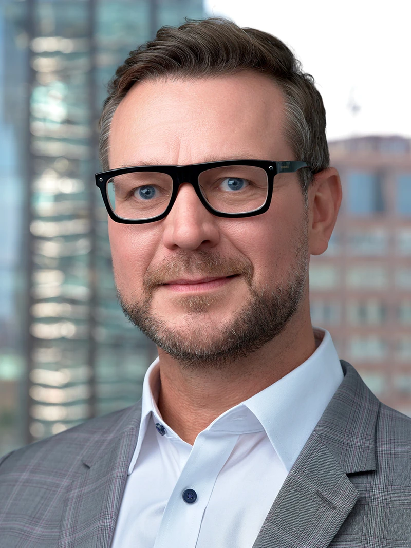 Corporate headshot with an office and city skyline background, a bearded man in glasses and a grey checked jacket, photographed on location