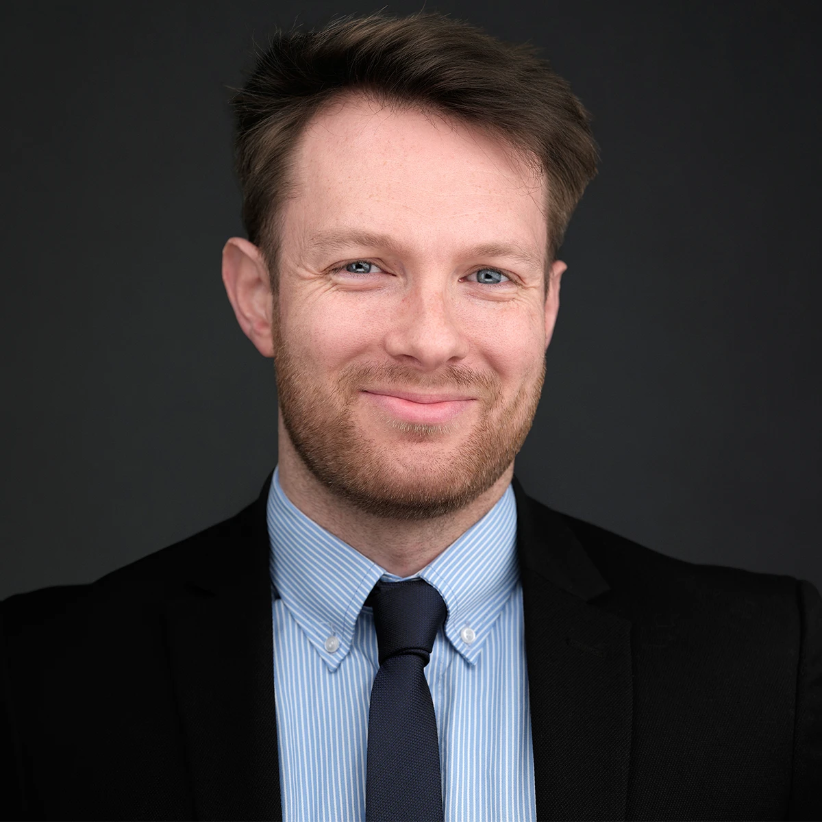 Corporate headshot of a man in a black suit and blue striped shirt, relaxed confident smile, dark studio background