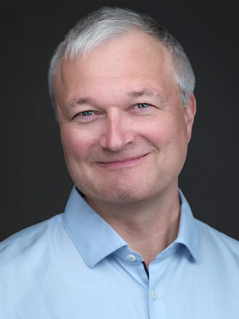 Corporate headshot on a dark background, a grey-haired man in a pale blue shirt with a warm, approachable smile