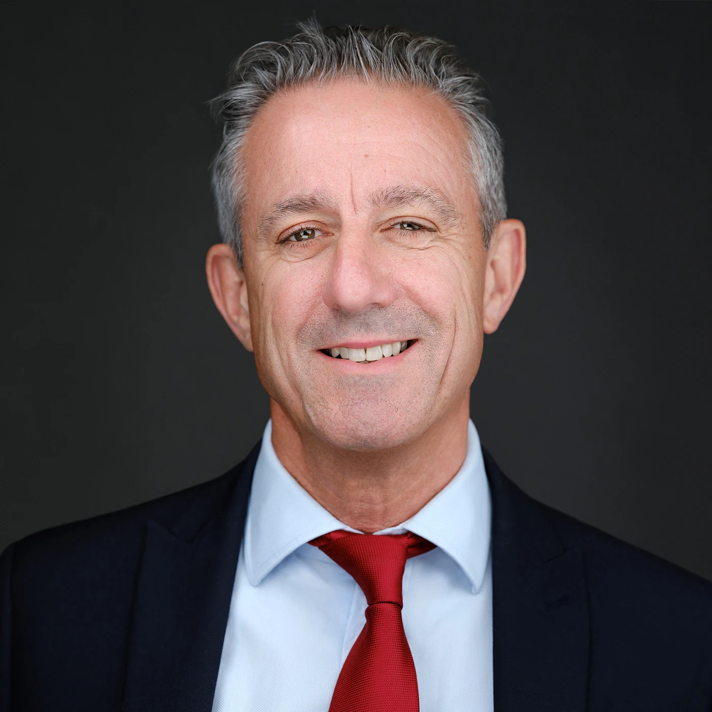 LinkedIn headshot of a smiling man with grey hair in a navy suit, light blue shirt and red tie on a dark background