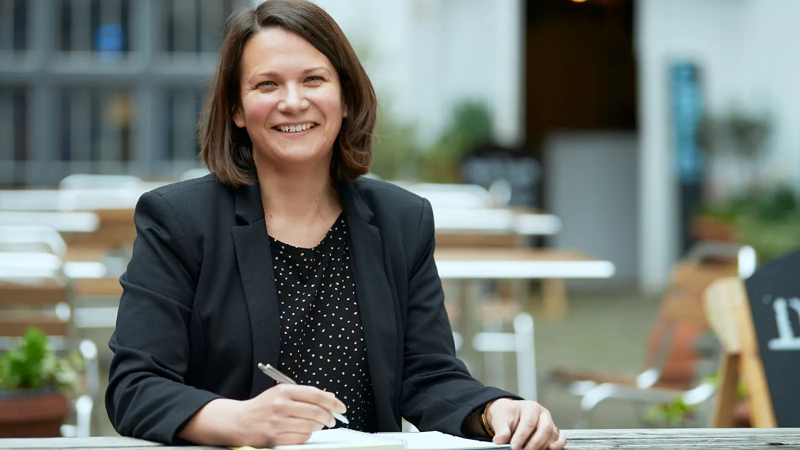 Woman in a black blazer smiling at an outdoor café table with a notebook
