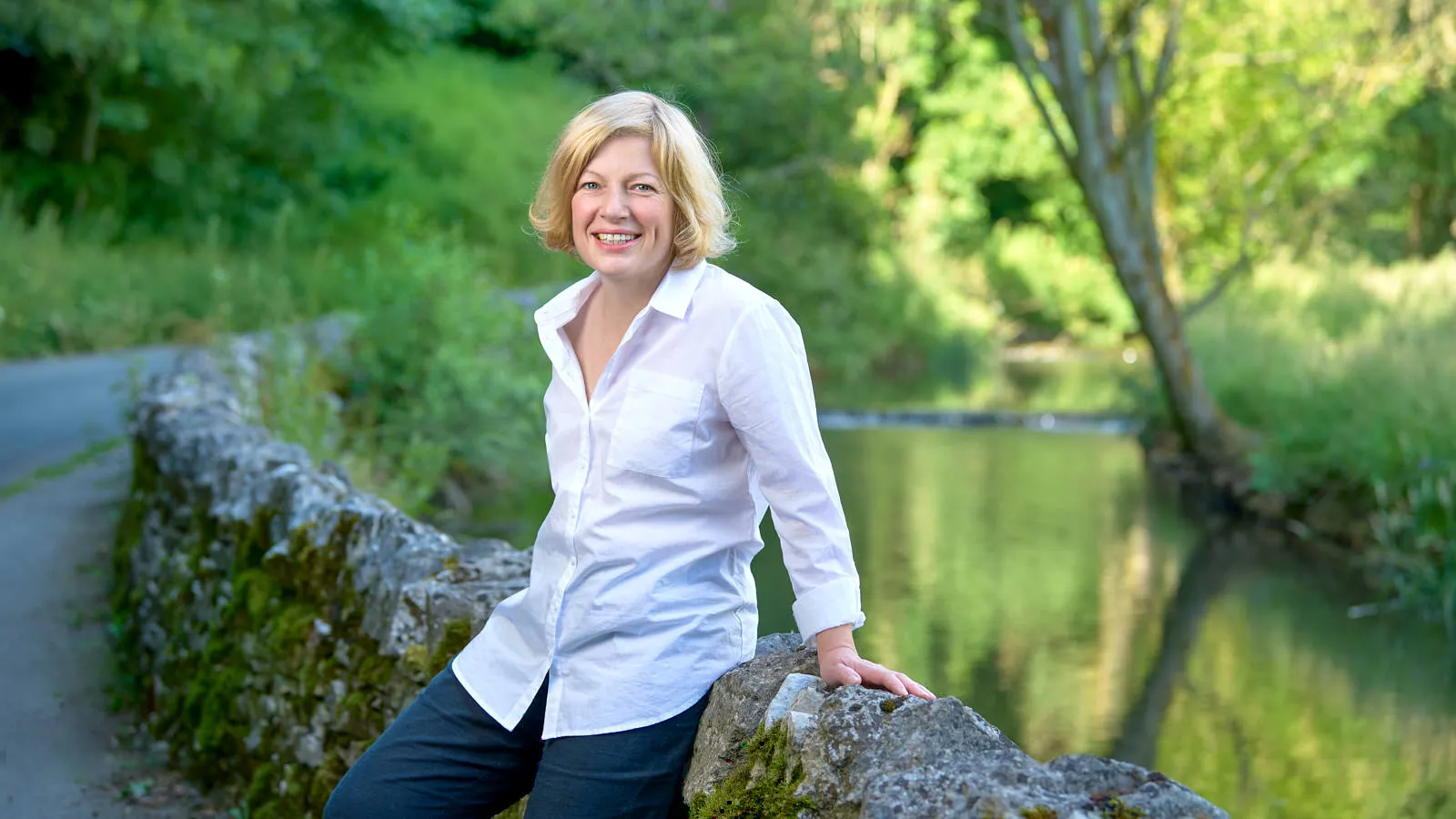 Woman in a white shirt smiling by a riverside stone wall, personal branding on location